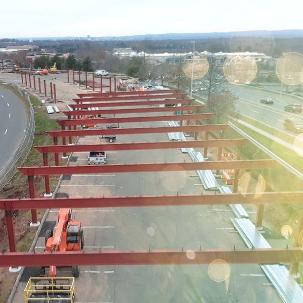 Aerial view of a solar carport steel frame under construction over a parking lot beside a highway
